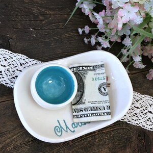 Pode incluir: Uma bandeja de cer&acirc;mica branca com uma tigela pequena e redonda de cor turquesa e uma nota de um d&oacute;lar dobrada. A palavra "Mara" est&aacute; escrita em script turquesa no prato. Flores decorativas e acabamento em renda no fundo.
