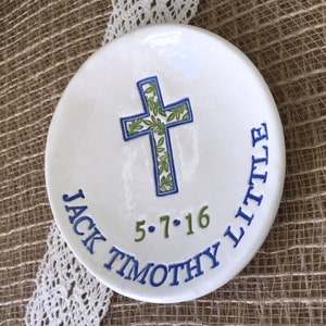 May include: White ceramic dish with a blue and green cross design. The cross is filled with green leaves and outlined in blue. The dish has the text "JACK TIMOTHY LITTLE" and the date "5.7.16" in blue and green.