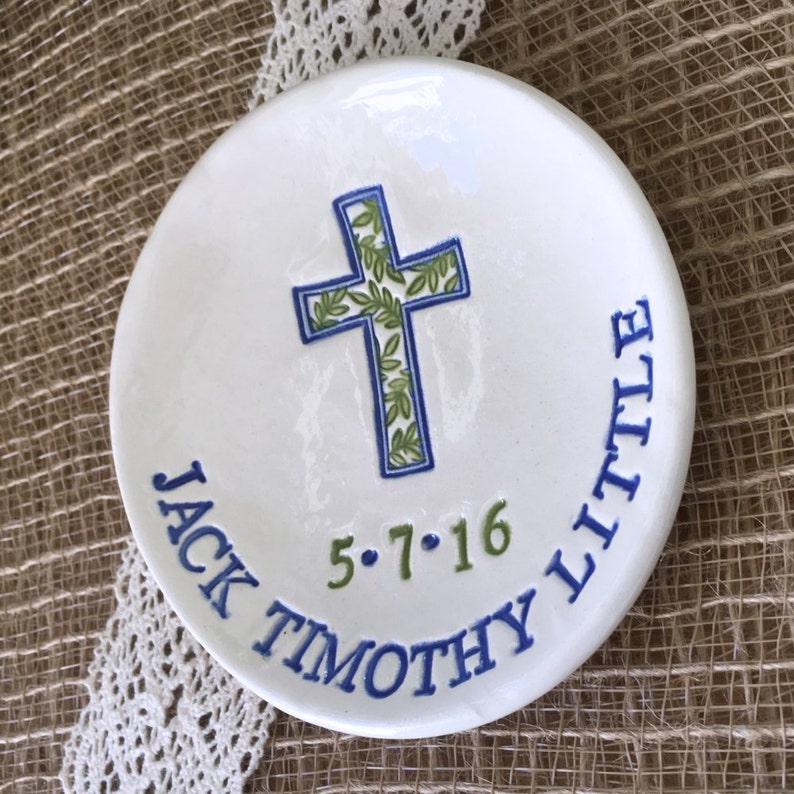 May include: White ceramic dish with a blue and green cross design. The cross is filled with green leaves and outlined in blue. The dish has the text "JACK TIMOTHY LITTLE" and the date "5.7.16" in blue and green.
