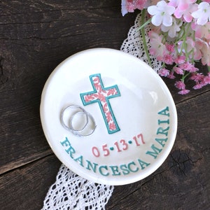 May include: White ceramic ring dish with a teal and coral cross design. The dish has the name "FRANCESCA MARIA" and the date "05-13-17" in teal and coral lettering. Two silver rings rest inside the dish. A wedding or anniversary keepsake.