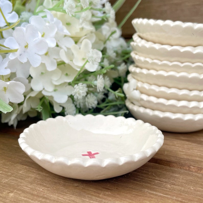 Puede incluir: Un conjunto de peque&ntilde;os cuencos de cer&aacute;mica blanquecina con bordes festoneados. Uno de los cuencos muestra un dise&ntilde;o de cruz rosa. Flores blancas en el fondo crean una est&eacute;tica suave y natural. Los cuencos est&aacute;n sobre una superficie de madera.