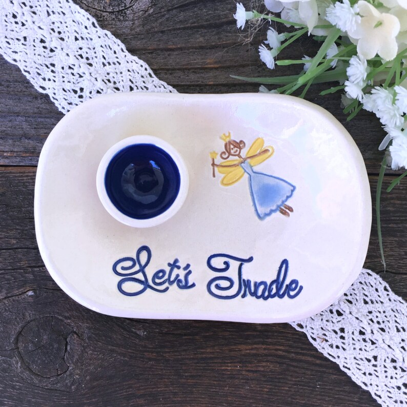 Pode incluir: Um prato de cer&acirc;mica branco oval com uma pequena tigela redonda azul escuro. O prato apresenta uma ilustra&ccedil;&atilde;o de fada e as palavras "Let's Trade" em escrita azul. Descansa sobre uma superf&iacute;cie de madeira escura.