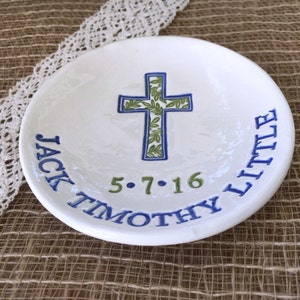 May include: A white ceramic dish with a blue and green cross design. The cross is decorated with green leaves and outlined in blue. The dish is inscribed with the name "JACK TIMOTHY LITTLE" and the date "5.7.16" in blue and green.