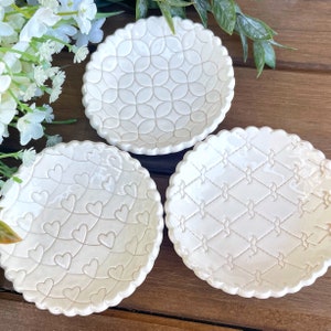 May include: Three off-white ceramic dishes with scalloped edges. Each dish features a different embossed pattern: interlocking circles, connected hearts, and a diamond lattice design. The dishes are displayed on a wooden surface.