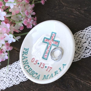 May include: White ceramic ring dish with a teal cross featuring a floral pattern in pink and white. The dish has the name "FRANCESCA MARIA" and the date "05.13.17" in teal and pink. Two silver rings rest on the dish.