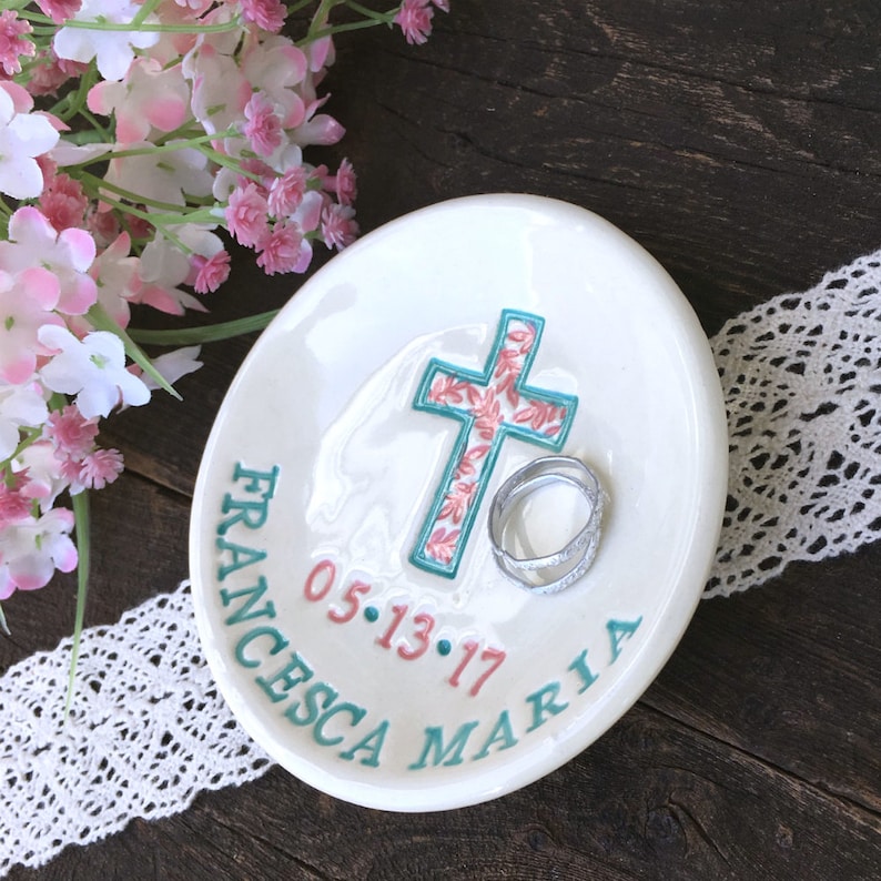 May include: White ceramic ring dish with a teal cross featuring a floral pattern in pink and white. The dish has the name "FRANCESCA MARIA" and the date "05.13.17" in teal and pink. Two silver rings rest on the dish.