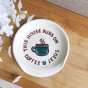 May include: White ceramic spoon rest with the text "THIS HOUSE RUNS ON COFFEE & JESUS" in brown lettering. A teal coffee cup graphic is in the center. The spoon rest is on a wooden surface.