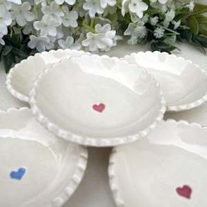 May include: A set of small, white ceramic dishes with a scalloped rim. Each dish has a hand-painted heart in red or blue. The dishes are arranged on a white surface, with white flowers in the background.