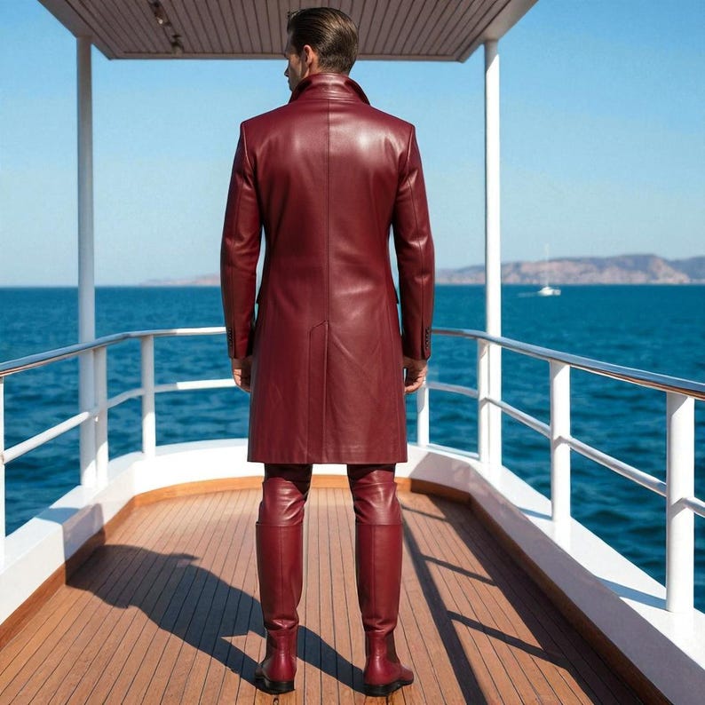 May include: A burgundy leather coat and matching pants are worn on a yacht. The long coat has a tailored fit and button details. The pants are fitted, and the boots are knee-high. The background shows the ocean and a clear blue sky.