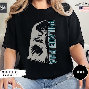 K&ouml;nnte beinhalten: Ein schwarzes T-Shirt mit einem Aufdruck eines gr&uuml;nen und grauen Adlerkopfes und dem Wort "PHILADELPHIA" in gr&uuml;nem Text. Das Shirt wird von einer Person getragen.