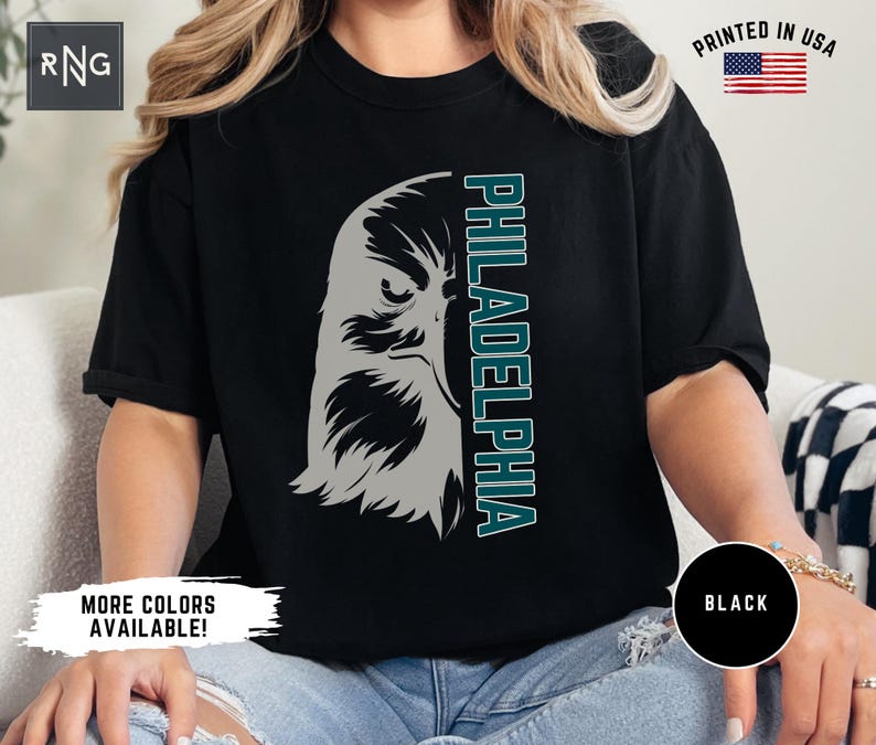 K&ouml;nnte beinhalten: Ein schwarzes T-Shirt mit einem Aufdruck eines gr&uuml;nen und grauen Adlerkopfes und dem Wort "PHILADELPHIA" in gr&uuml;nem Text. Das Shirt wird von einer Person getragen.