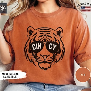 Könnte beinhalten: Ein rostfarbenes Kurzarm-T-Shirt mit einer schwarzen Grafik eines Tigers, der eine Sonnenbrille trägt. Auf der Sonnenbrille steht in weißer Schrift CIN CY. Das T-Shirt hat einen Rundhalsausschnitt und eine lockere Passform.