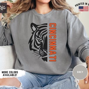 Puede incluir: Una sudadera gris con un gr&aacute;fico de cabeza de tigre en blanco y negro y la palabra "CINCINNATI" en letras naranjas.