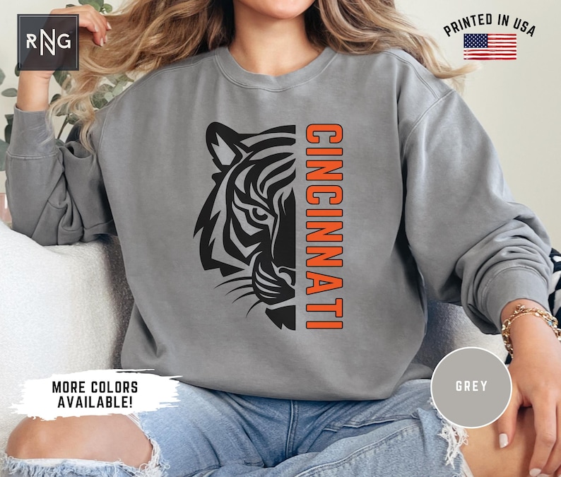Puede incluir: Una sudadera gris con un gr&aacute;fico de cabeza de tigre en blanco y negro y la palabra "CINCINNATI" en letras naranjas.
