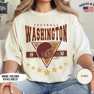 May include: An ivory short-sleeve t-shirt featuring a graphic print with the text WASHINGTON FOOTBALL and the numbers 1932. The design includes a maroon football helmet centred over a yellow triangle, accented by yellow stars and horizontal lines.