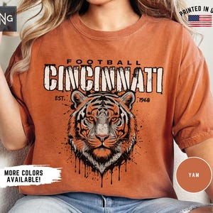 May include: A rust-colored short-sleeve t-shirt featuring a graphic of a tiger face with black ink-splatter details. The text reads FOOTBALL CINCINNATI EST. 1968. The shirt includes a small logo in the top left corner and a printed in USA flag icon.