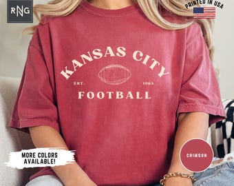 May include: Crimson-colored t-shirt with the text "Kansas City Football" in a curved arch, featuring a football graphic. The shirt includes the text "EST. 1963" and "Printed in USA". Additional text reads "More Colors Available!"