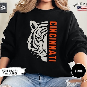 May include: A black long-sleeve sweatshirt featuring a white graphic of a tiger face on the left side and the word CINCINNATI in orange vertical text on the right. The design includes a small logo in the top left corner and a flag icon.