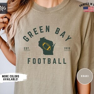 Peut inclure: Un t-shirt kaki avec un texte vert fonc&eacute; indiquant GREEN BAY FOOTBALL et EST. 1919. Le motif comprend une silhouette vert fonc&eacute; de l'&Eacute;tat du Wisconsin avec un graphique de ballon de football jaune et blanc centr&eacute; au milieu de l'&Eacute;tat.