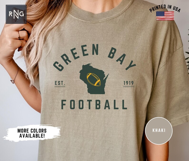 Peut inclure: Un t-shirt kaki avec un texte vert fonc&eacute; indiquant GREEN BAY FOOTBALL et EST. 1919. Le motif comprend une silhouette vert fonc&eacute; de l'&Eacute;tat du Wisconsin avec un graphique de ballon de football jaune et blanc centr&eacute; au milieu de l'&Eacute;tat.