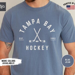 May include: A blue jean t-shirt with a white graphic of crossed hockey sticks and the text "Tampa Bay" in a white arc above the sticks. The text "Est. 1992" is below the sticks and the text "Hockey" is below that. The text "Printed in USA" is in the upper right corner of the image.