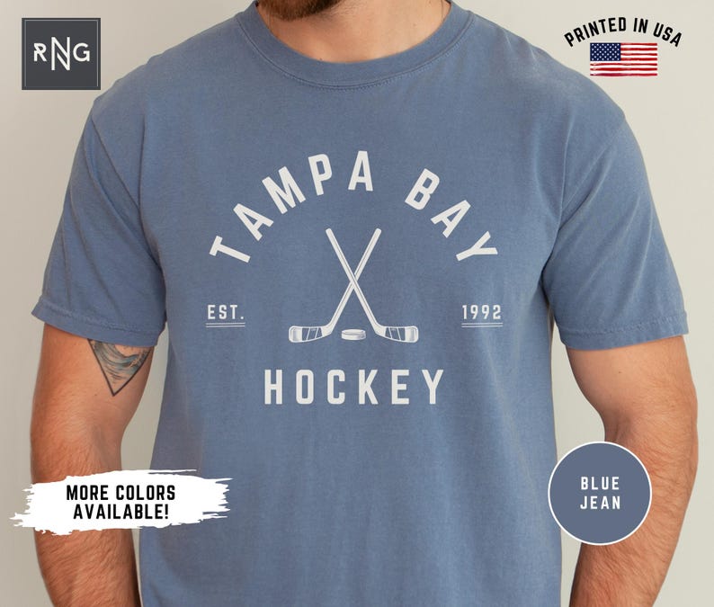 May include: A blue jean t-shirt with a white graphic of crossed hockey sticks and the text "Tampa Bay" in a white arc above the sticks. The text "Est. 1992" is below the sticks and the text "Hockey" is below that. The text "Printed in USA" is in the upper right corner of the image.