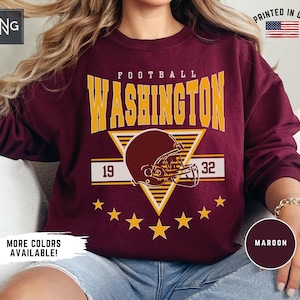 May include: A maroon long-sleeve crewneck sweatshirt featuring a yellow graphic print that reads FOOTBALL WASHINGTON with the numbers 19 and 32 on either side of a football helmet illustration, accented by a series of yellow stars at the bottom.