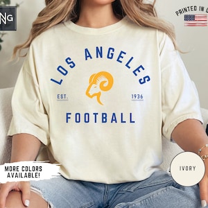 May include: A white t-shirt featuring a blue and yellow graphic of the Los Angeles Rams logo. The graphic includes the text "Los Angeles" in a blue arc above the logo, "Est. 1936" below the logo, and "Football" below the text. The t-shirt is being modelled by someone wearing blue jeans.