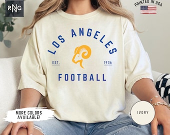 Op de afbeelding: Een wit T-shirt met een blauwe en gele afbeelding van het Los Angeles Rams logo. De afbeelding bevat de tekst "Los Angeles" in een blauwe boog boven het logo, "Est. 1936" onder het logo en "Football" onder de tekst. Het T-shirt wordt gedragen door een model dat een blauwe jeans draagt.