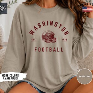 May include: A sandstone-coloured long-sleeve crewneck sweatshirt featuring the maroon text WASHINGTON FOOTBALL, a graphic of a football helmet, and the text EST. 1932. The design is centred on the chest of the garment.
