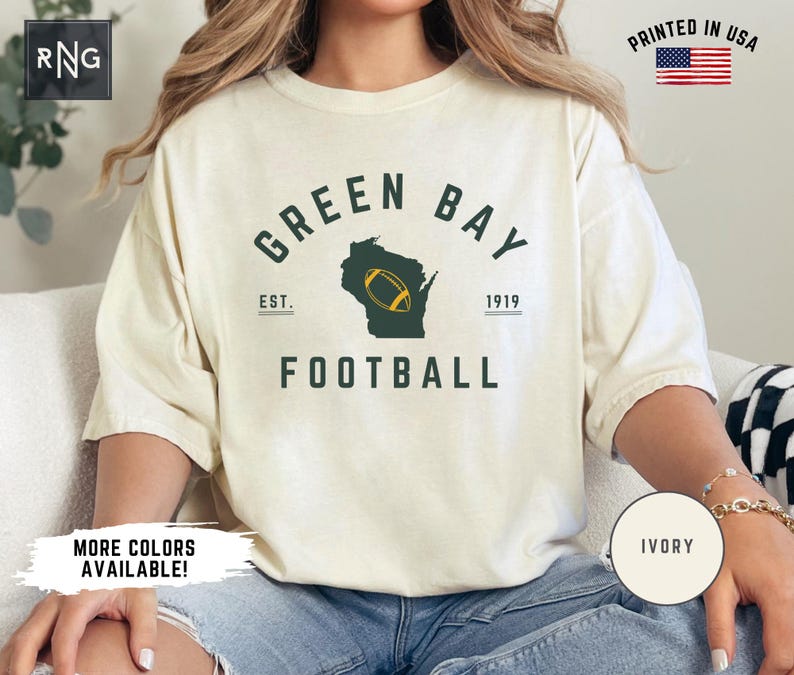 Peut inclure: Un t-shirt &agrave; manches courtes couleur ivoire avec le texte vert fonc&eacute; GREEN BAY FOOTBALL et EST. 1919 de chaque c&ocirc;t&eacute;. Une silhouette vert fonc&eacute; de l'&Eacute;tat du Wisconsin est centr&eacute;e avec un graphique de ballon de football jaune et blanc.