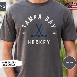 May include: A dark gray t-shirt with a white graphic of the words "Tampa Bay Hockey" with a hockey stick and puck design. The text "Est. 1992" is also included in the graphic.