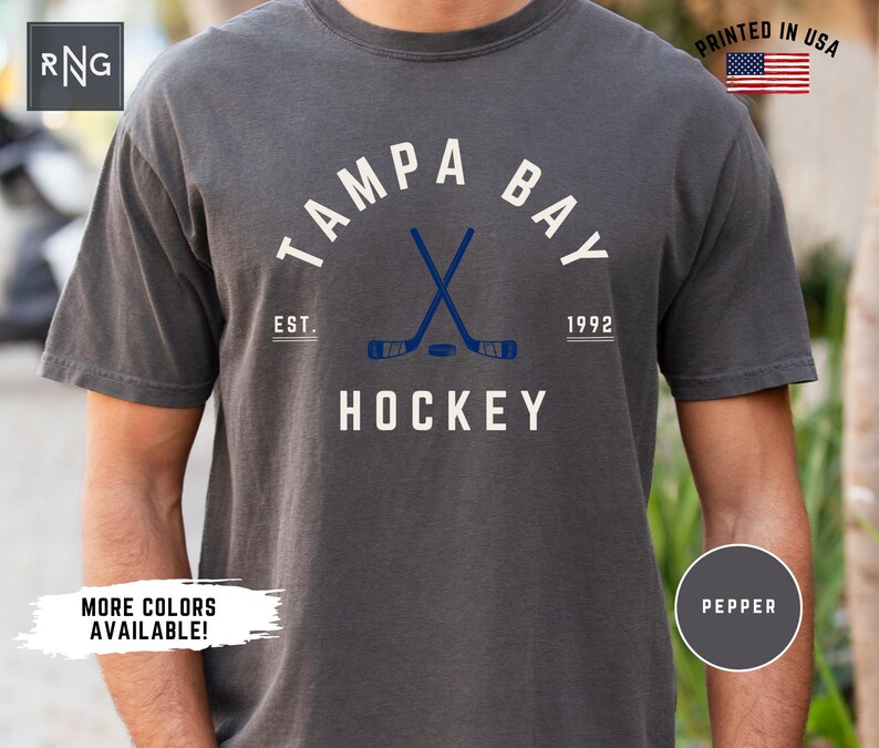 May include: A dark gray t-shirt with a white graphic of the words "Tampa Bay Hockey" with a hockey stick and puck design. The text "Est. 1992" is also included in the graphic.