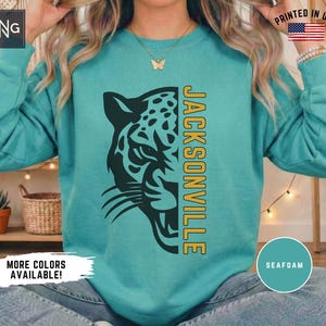 May include: A seafoam green long-sleeve sweatshirt featuring a dark green graphic of a jaguar head on the left side and the word JACKSONVILLE printed vertically in yellow on the right side. The garment has a crew neckline and long sleeves.
