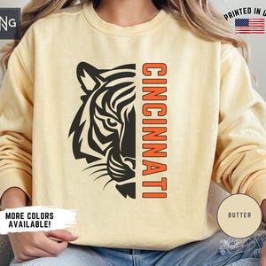 Puede incluir: Una sudadera amarilla con una cabeza de tigre negra y la palabra "CINCINNATI" en naranja en la parte delantera. La sudadera tiene una etiqueta "BUTTER" en la manga.