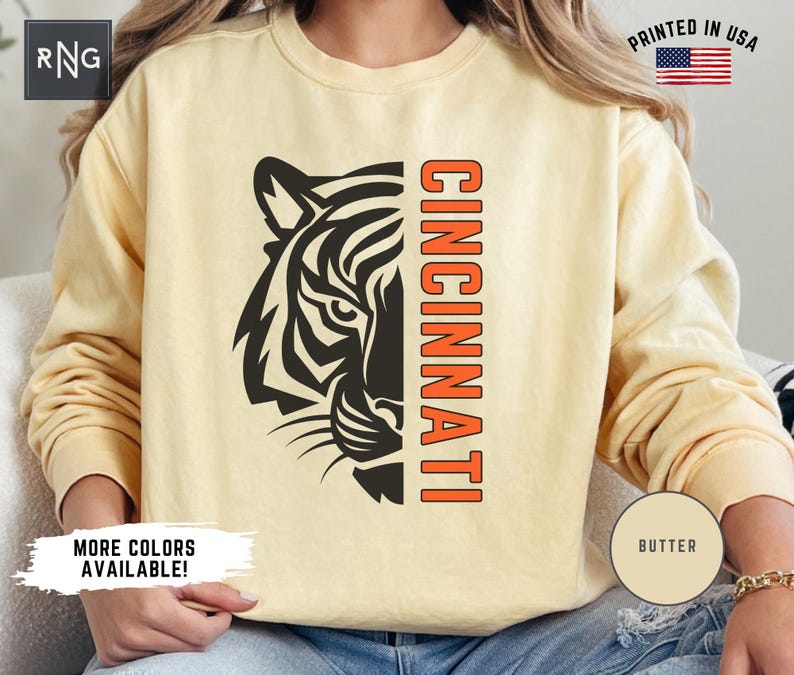Puede incluir: Una sudadera amarilla con una cabeza de tigre negra y la palabra "CINCINNATI" en naranja en la parte delantera. La sudadera tiene una etiqueta "BUTTER" en la manga.