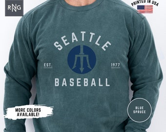 May include: A teal blue long-sleeved sweatshirt with a white graphic print of a baseball team logo. The logo features the text "Seattle Baseball" with the team's mascot, a trident, in a blue circle. The text "Est. 1977" is printed below the logo.