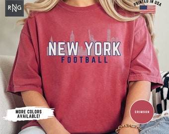 May include: A crimson short-sleeve t-shirt featuring a graphic of the New York City skyline in light grey. The text NEW YORK is printed in large white letters with a dark blue outline, and the word FOOTBALL is printed below in dark blue.