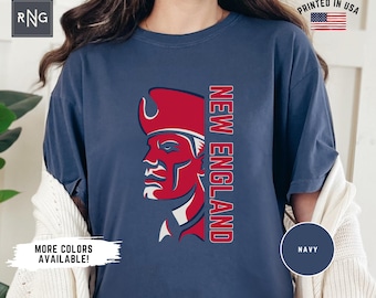May include: Navy blue t-shirt featuring a red and white graphic of a colonial figure with the text "NEW ENGLAND" in red. The shirt has a relaxed fit and is printed in the USA. Additional colors are available.