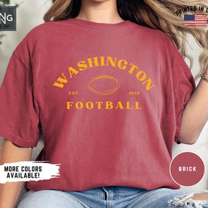 May include: A brick-red t-shirt with the words "WASHINGTON FOOTBALL" in yellow, and a football graphic. The shirt also includes "EST. 1932". The words "MORE COLORS AVAILABLE!" are visible.