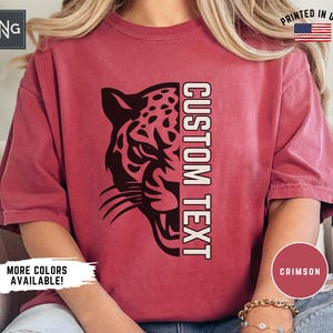 May include: Crimson-colored t-shirt featuring a black leopard graphic on the left side and the words "CUSTOM TEXT" in white on the right. The shirt has short sleeves and a relaxed fit. The text "MORE COLORS AVAILABLE!" is also visible.