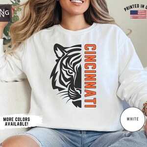 Puede incluir: Sudadera blanca con un gr&aacute;fico de tigre blanco y negro en el lado izquierdo y la palabra "CINCINNATI" en naranja en el lado derecho.