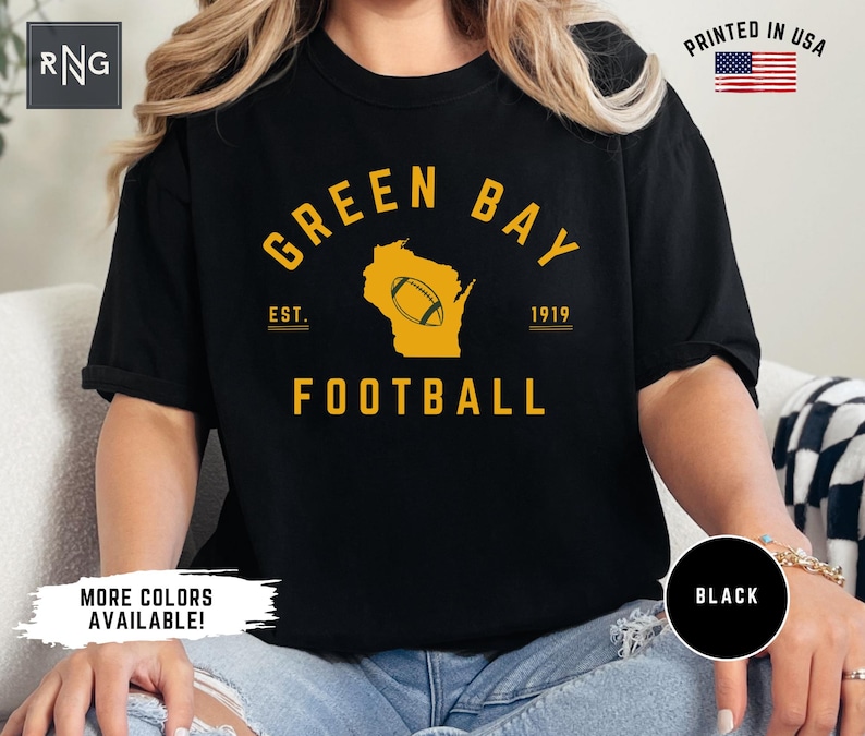 Peut inclure: Un t-shirt noir &agrave; manches courtes avec un imprim&eacute; graphique jaune indiquant GREEN BAY FOOTBALL, EST. 1919 et une silhouette de l'&Eacute;tat du Wisconsin contenant une ic&ocirc;ne de ballon de football. Le t-shirt porte un logo RNG et un drapeau PRINTED IN USA.
