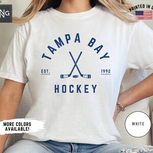 May include: White t-shirt with a blue graphic of crossed hockey sticks and a puck. The graphic includes the text "Tampa Bay" in a semi-circle above the sticks and "EST. 1992" below the sticks. The text "HOCKEY" is below the sticks and puck.