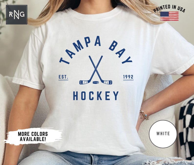 May include: White t-shirt with a blue graphic of crossed hockey sticks and a puck. The graphic includes the text "Tampa Bay" in a semi-circle above the sticks and "EST. 1992" below the sticks. The text "HOCKEY" is below the sticks and puck.