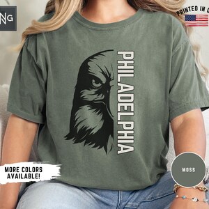 K&ouml;nnte beinhalten: Ein gr&uuml;nes T-Shirt mit einem schwarzen und wei&szlig;en Adler-Motiv und dem Text "PHILADELPHIA" auf der Vorderseite. Das Shirt ist aus einem weichen, bequemen Material gefertigt und hat eine lockere Passform.