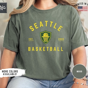 May include: Green t-shirt with a yellow and green Seattle Basketball logo. The logo features a basketball in a net and a shield. The text "Seattle" is in a yellow arch above the logo. The text "Est. 2000" is below the logo. The text "Basketball" is in yellow below the logo.