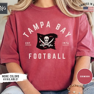 Puede incluir: Una camiseta de color carmesí con un gráfico blanco de una bandera pirata con una calavera y huesos cruzados. El texto "Tampa Bay" está por encima de la bandera y "EST. 1976" está por debajo de la bandera. El texto "FOOTBALL" está por debajo de la bandera. El texto "MORE COLORS AVAILABLE!" está en la esquina inferior izquierda de la imagen. El texto "CRIMSON" está en la esquina inferior derecha de la imagen.