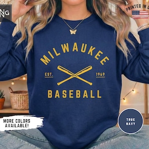 May include: A navy blue crewneck sweatshirt featuring yellow text that reads MILWAUKEE BASEBALL with EST. 1969 and a graphic of two crossed baseball bats in the center. The design is printed on the front of the long-sleeved pullover garment.