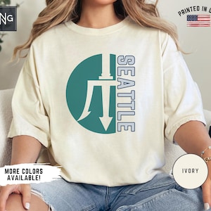 May include: A white t-shirt with a teal circle and a white trident graphic. The text "SEATTLE" is printed vertically in blue on the right side of the circle. The shirt is available in ivory.